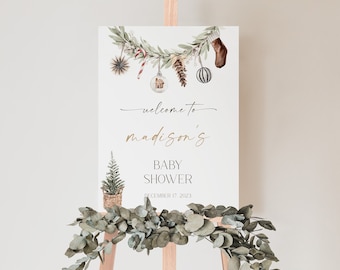 May include: A white sign on a wooden easel, decorated with a watercolor garland of greenery and Christmas ornaments. The sign reads "welcome to madison's BABY SHOWER December 17 2023". A small Christmas tree in a woven basket is also featured.