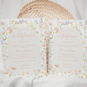 May include: Two baby shower invitations with a floral design. The invitations are white with pink and blue flowers and butterflies. The text on the invitations reads "Our Little Wildflower is almost here!" and includes the baby shower details.