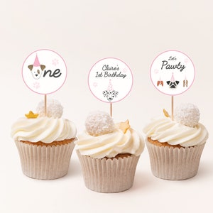 May include: Three cupcakes with white frosting and pink and white dog-themed cupcake toppers. The toppers say "One", "Claire's 1st Birthday", and "Let's Pawty".