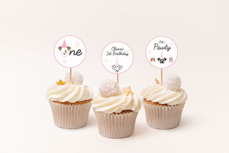 May include: Three cupcakes with white frosting and pink and white dog-themed cupcake toppers. The toppers say "One", "Claire's 1st Birthday", and "Let's Pawty".
