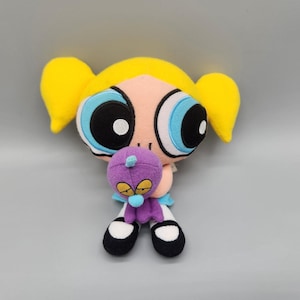 May include: A plush doll of the Powerpuff Girls character Bubbles, with yellow pigtails, large blue eyes, and a pink face. She is holding a small purple plush toy with yellow eyes and a blue nose. The doll is wearing a blue dress and black and white shoes.