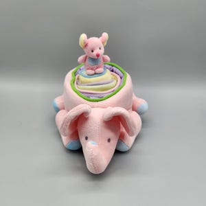 May include: A soft pink plush elephant toy with blue accents on its feet and ears. A smaller pink mouse toy sits atop a colorful, layered structure inside the elephant's back. The layers are pastel colors.