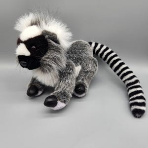 May include: A plush lemur toy with a black and white striped tail. The lemur has a black face, white muzzle, and a fluffy gray and white body. The toy is made of soft, plush material and has black paws.