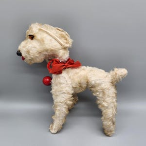 May include: A vintage stuffed dog toy with a cream-colored, shaggy fur texture. It has a red bow around its neck and a red ball hanging below. The dog has brown eyes, a black nose, and red lips. The toy is standing on all fours.