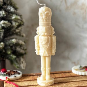 Può includere: Candela color avorio a forma di soldato schiaccianoci. La candela è posizionata su una slitta di legno con elementi decorativi, tra cui un piccolo albero di Natale e ornamenti rotondi. La candela presenta dettagli intricati, tra cui un cappello alto e un'uniforme.