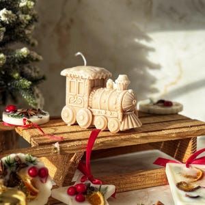 Peut inclure: Une bougie beige en forme de train est posée sur une luge en bois, entourée de bougies en forme d'étoile et rondes décorées de baies rouges, de tranches d'orange et de verdure. Une petite cloche dorée est au premier plan. Thème de vacances.
