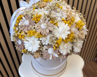 May include: A round floral arrangement in a white box, featuring white, tan, and yellow flowers. The flowers are arranged in a dense, compact cluster, with a white ribbon tied on top. The box has a gold trim and a rope detail.