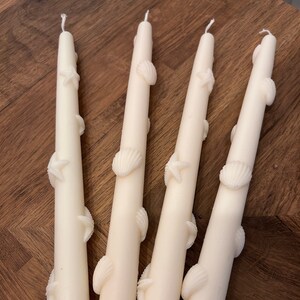 May include: Four white taper candles with seashell and starfish designs. The candles are arranged in a row on a wooden surface.