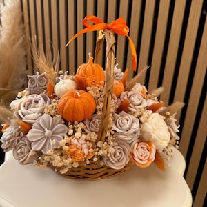 Puede incluir: Una cesta decorativa rebosante de elementos temáticos de otoño. Presenta velas con forma de calabaza naranja, arreglos florales de jabón gris y blanco y una cinta naranja. La cesta está llena de flores secas y una figura de búho.
