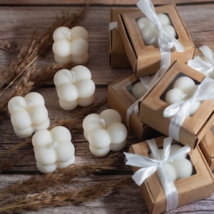 May include: White, round, multi-faceted candles in small, brown boxes tied with white ribbon. The boxes are stacked on a wooden surface with dried grass.
