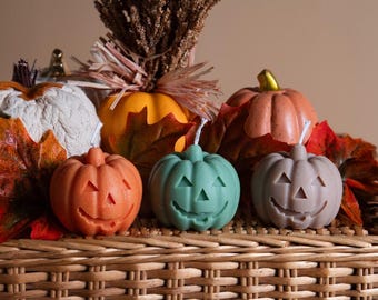Könnte beinhalten: Drei Halloween-Kürbiskerzen in Orange, Grün und Grau, die auf einem geflochtenen Weidenkorb mit Herbstlaub liegen.