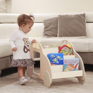 Può includere: Un carrello porta libri in legno bianco a forma di auto con ruote. Lo scaffale ha tre ripiani per i libri. Un bambino piccolo è in piedi accanto allo scaffale.
