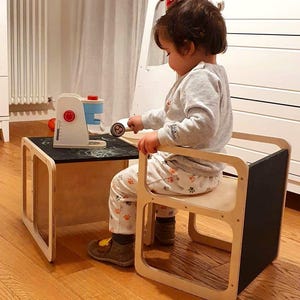 Può includere: Un bambino è seduto a un set di tavolo e sedia in legno con un piano in ardesia nero. Il tavolo e la sedia sono entrambi in legno chiaro con una finitura naturale. Il bambino sta giocando con una macchina per il caffè giocattolo.