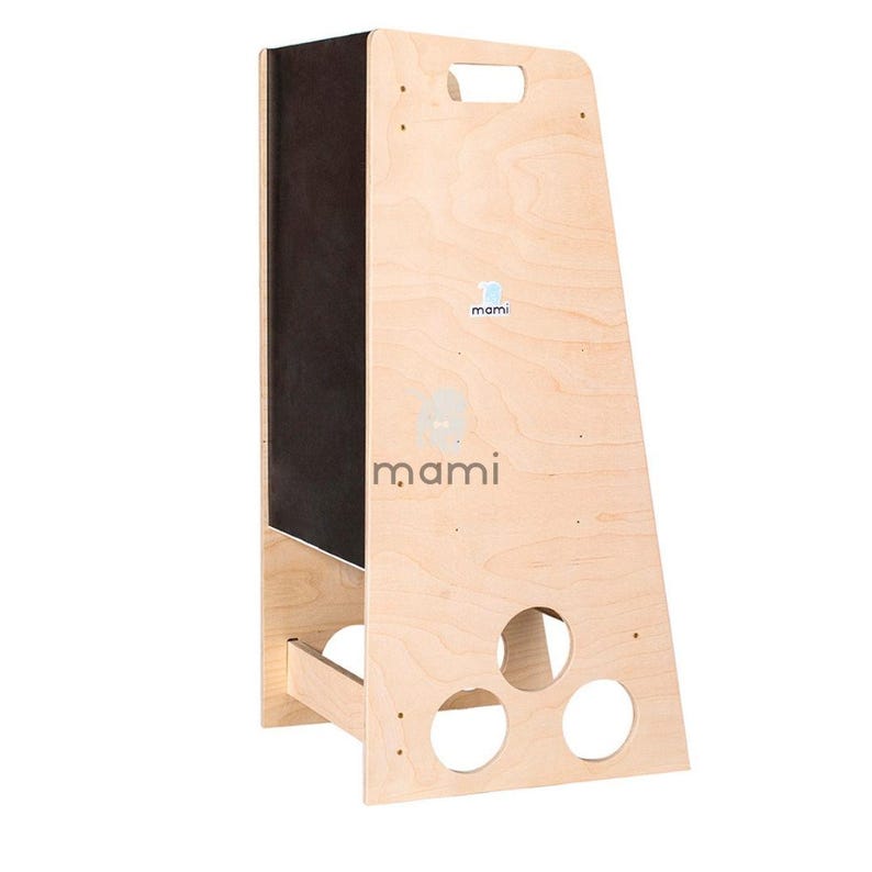 Puede incluir: Un taburete de madera con un panel de pizarra negra en un lado. El taburete tiene un asa recortada y tres agujeros redondos en la parte inferior. El taburete est&aacute; etiquetado con "mami" en ambos lados.