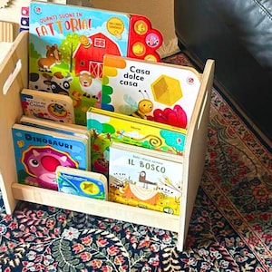 Può includere: Una libreria in legno piena di libri per bambini colorati. I libri hanno titoli in italiano, tra cui "Quanti Suoni in Fattoria", "Casa Dolce Casa", "Il Bosco" e "Dinosauri".