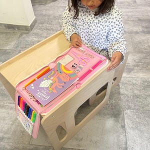 Può includere: Un tavolo in legno rosa e bianco con un set di arte a tema unicorno rosa e bianco. Il set include un libro da colorare, matite colorate, pennarelli e altri materiali artistici. Il tavolo ha un vano portaoggetti integrato per i materiali artistici.