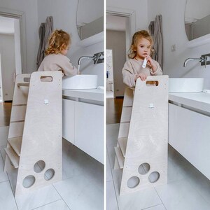 Puede incluir: Un taburete de madera blanco con un asa y tres c&iacute;rculos recortados. Un ni&ntilde;o est&aacute; de pie en el taburete y se cepilla los dientes en un lavabo blanco.