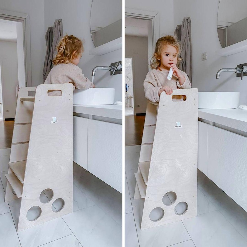 Puede incluir: Un taburete de madera blanco con un asa y tres c&iacute;rculos recortados. Un ni&ntilde;o est&aacute; de pie en el taburete y se cepilla los dientes en un lavabo blanco.