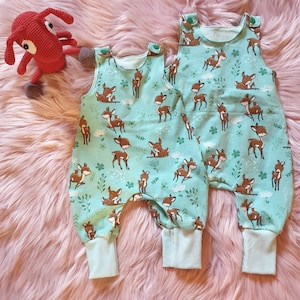 Könnte beinhalten: Zwei Paar Baby-Latzhosen mit hellblauem Hintergrund und einem braunen Hirsch-Druck. Die Latzhosen haben einen grünen Knopf an jeder Schultertrage.