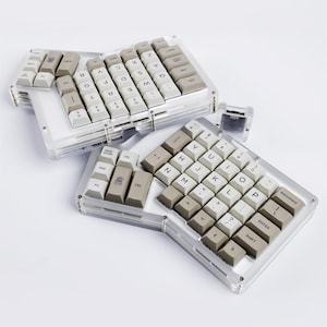 May include: A split ergonomic mechanical keyboard with a clear acrylic case and beige keycaps. The layout is divided into two separate sections, each featuring a grid of keys with black lettering and a small cactus icon on one key.
