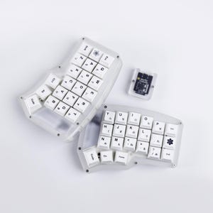 May include: A split ergonomic mechanical keyboard with a translucent frosted case and white keycaps featuring black Latin letters and Japanese hiragana characters. The keyboard includes a small separate square component with a black circuit board.