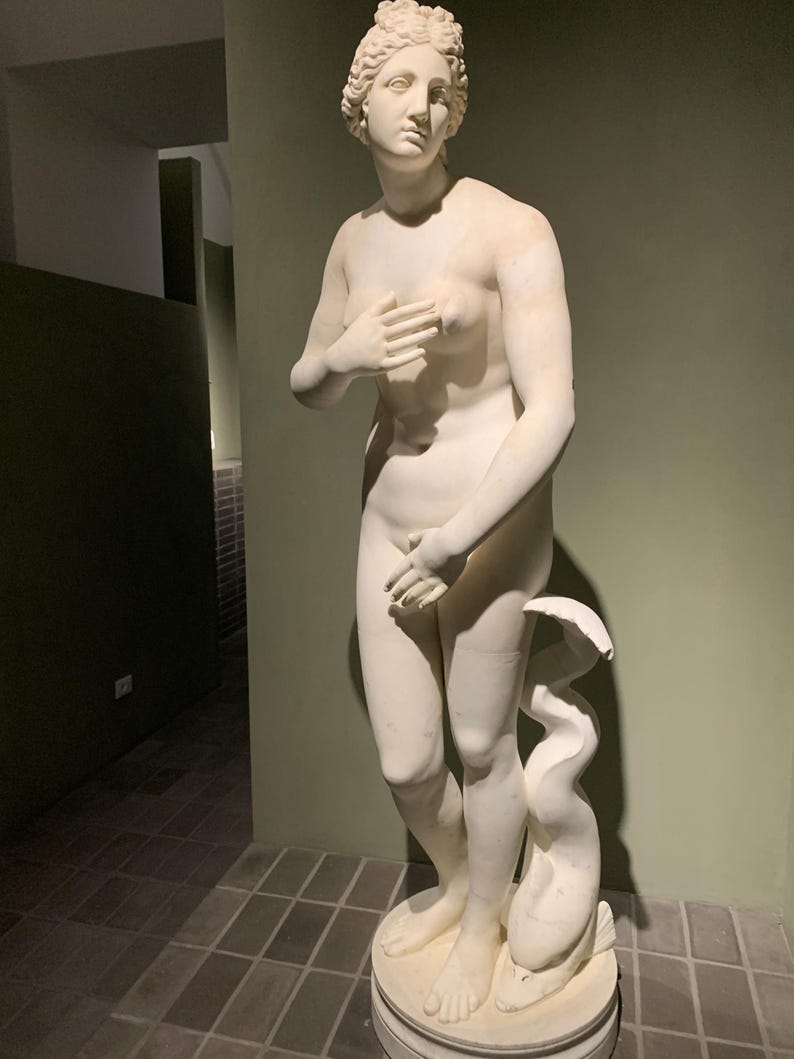 May include: A white marble statue of a woman standing with her right hand on her chest and her left hand on her hip. She is wearing a simple, flowing robe and has a serene expression on her face. The statue is standing on a pedestal with a dolphin at her feet.