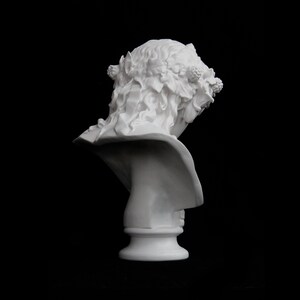 May include: White plaster bust sculpture of a person with a wreath of leaves and berries on their head. The bust is facing away from the viewer and is on a white pedestal.