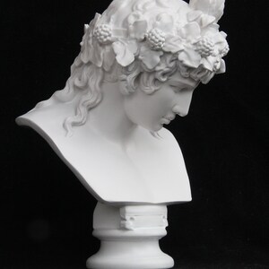 May include: White plaster bust of a person with a floral crown. The bust is on a white pedestal.