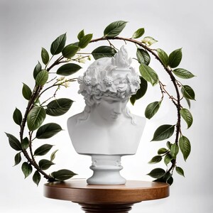 May include: White plaster bust of a person with a wreath of leaves on their head, set on a wooden table with a green leafy garland around it.