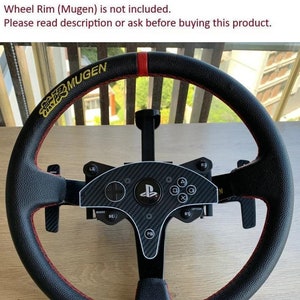 May include: Black steering wheel with red stitching and a carbon fiber center console. The wheel has the text "MUGEN" and PlayStation controller buttons. The image also includes the text "Wheel Rim (Mugen) is not included."