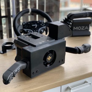 May include: Black sim racing equipment, including a steering wheel, a control box with a fan, and a gear shift lever. The control box has the brand name "MOZA" printed on it. The items are on a light wood surface.