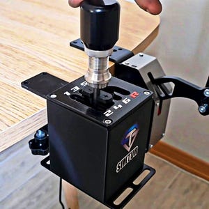 Könnte beinhalten: Ein schwarzer und silberner Rennsimulator-Schalthebel mit schwarzem Griff. Der Schalthebel hat Ganganzeigen und das Logo "Simtur". Das Gerät ist an einer schwarzen Halterung befestigt und an einer Holzoberfläche angebracht.