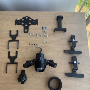 Op de afbeelding: Een set zwarte kunststof onderdelen voor een race-simulator, inclusief een stuurwiel, pedalen en een versnellingspook. De onderdelen liggen op een houten tafel.