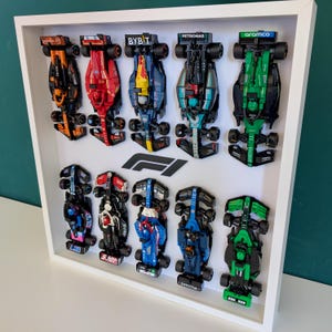 Peut inclure: Un présentoir encadré blanc avec douze voitures de course miniatures de Formule 1. Les voitures sont de différentes couleurs, notamment orange, rouge, bleu, noir et vert, et sont disposées en deux rangées. Le logo F1 est au centre.