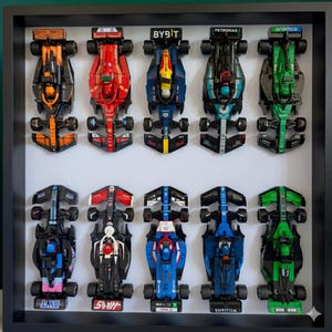 Op de afbeelding: Een ingelijste verzameling van tien kleurrijke speelgoed raceauto's, elk met unieke ontwerpen en sponsorlogo's. De auto's zijn gerangschikt in twee rijen van vijf, met een verscheidenheid aan kleuren, waaronder rood, groen, blauw en zwart. De lijst is zwart en de achtergrond is wit.