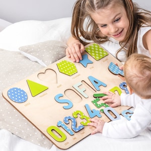 Puede incluir: Un tablero de rompecabezas de madera con formas, letras y números coloridos. Las formas incluyen un círculo, un triángulo, un corazón, un hexágono y una estrella. Las letras deletrean el nombre "SOFIA". Los números son del 0 al 7. Las piezas del rompecabezas tienen un patrón de lunares azules y blancos.