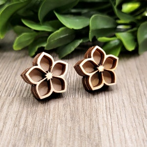 K&ouml;nnte beinhalten: Ein Paar h&ouml;lzerne Ohrstecker in Blumenform. Die Ohrringe bestehen aus geschichtetem Holz mit einer hellbraunen Mitte und dunkleren braunen R&auml;ndern. Die Ohrringe sind vor einem verschwommenen Hintergrund aus gr&uuml;nen Bl&auml;ttern und einer Holzoberfl&auml;che platziert.