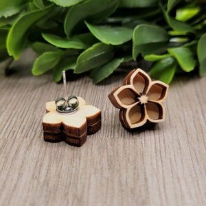 K&ouml;nnte beinhalten: Ohrstecker in Form einer Holzblume. Die Ohrringe bestehen aus geschichtetem Holz mit einer hellbraunen Mitte und dunkleren braunen R&auml;ndern, wodurch ein dreidimensionaler Effekt entsteht. Die Ohrringpfosten sind aus silberfarbenem Metall.