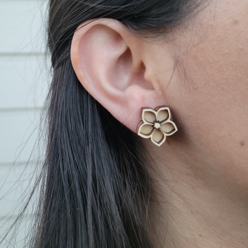 K&ouml;nnte beinhalten: Ohrstecker aus Holz in Blumenform. Die Ohrringe sind hellbraun mit einer dunkleren braunen Kontur und haben die Form einer f&uuml;nfbl&auml;ttrigen Blume. Die Ohrringe werden am Ohr getragen.