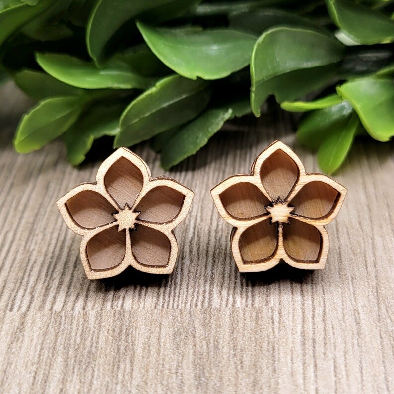 K&ouml;nnte beinhalten: Zwei h&ouml;lzerne Ohrstecker in Blumenform. Jeder Ohrring hat ein f&uuml;nfbl&auml;ttriges Design mit einem sternf&ouml;rmigen Zentrum. Die Ohrringe sind hellbraun und liegen auf einer hellen Holzmaserungsoberfl&auml;che, mit gr&uuml;nem Laub im Hintergrund.