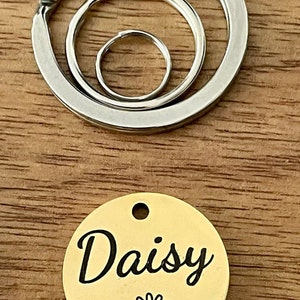 May include: Gold-colored metal pet tag with the name "Daisy" engraved in black cursive lettering and a black daisy graphic. The tag is attached to a silver key ring with three interlocking rings.