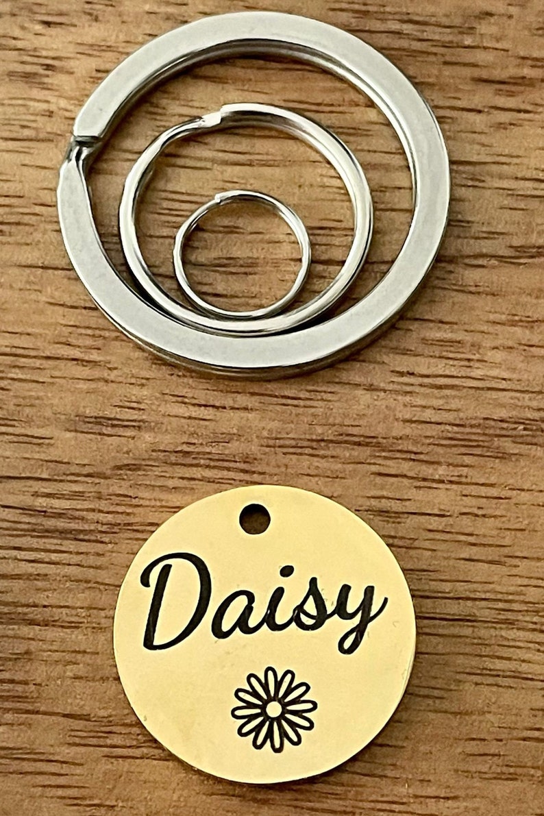 May include: Gold-colored metal pet tag with the name "Daisy" engraved in black cursive lettering and a black daisy graphic. The tag is attached to a silver key ring with three interlocking rings.