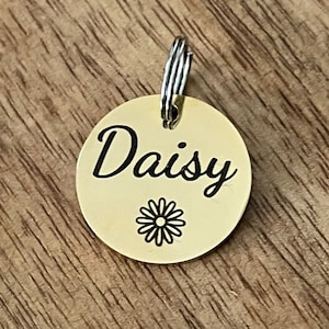May include: Gold-colored round pet tag with the name "Daisy" engraved in black cursive lettering and a small black daisy graphic.