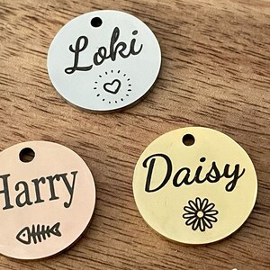 May include: Three round metal pet ID tags with different names and designs. The first tag is silver with the name "Loki" and a heart design. The second tag is rose gold with the name "Harry" and a fishbone design. The third tag is gold with the name "Daisy" and a daisy design.