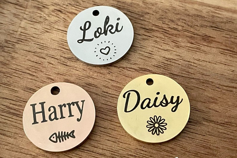 May include: Three round metal pet ID tags with different names and designs. The first tag is silver with the name "Loki" and a heart design. The second tag is rose gold with the name "Harry" and a fishbone design. The third tag is gold with the name "Daisy" and a daisy design.