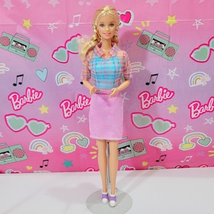 May include: A Barbie doll with blonde hair, wearing a plaid shirt, a pink skirt, and purple shoes. The doll is standing in front of a pink backdrop with the word "Barbie" and other graphics.