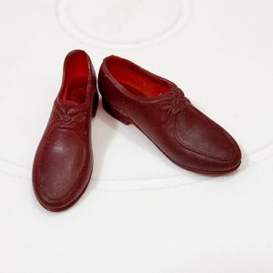May include: Two miniature burgundy-colored shoes with a classic design. The shoes have a rounded toe, a decorative bow detail, and a low heel. The interior of the shoes is a lighter shade of red.