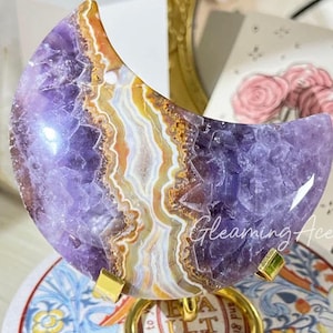 Puede incluir: Un colgante de ágata amatista en forma de media luna con un soporte dorado. El ágata tiene una base púrpura y blanca con remolinos naranja y amarillo. El soporte tiene las palabras "Mejores deseos para mi belleza" grabadas.