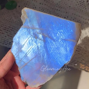 以下が含まれることがあります： 鮮やかな青色の虹色の光沢を持つ、研磨された生のムーンストーン。 石は自然な、ざらざらしたエッジと滑らかで反射する表面を持っています。 テキスト「Gleaming Ace」が石に表示されています。