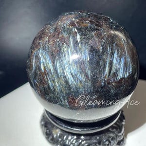 Puede incluir: Una esfera negra con vetas iridiscentes azules y marrones, descansando sobre un soporte negro con detalles intrincados. La esfera está hecha de un material natural, posiblemente una gema o un cristal.